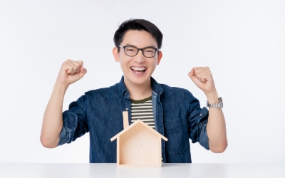 Seorang pria tersenyum sambil mengangkat kedua tangannya dengan miniatur rumah di depannya (freepik.com)